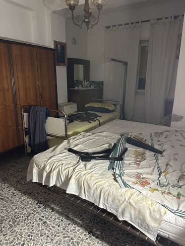 Es handelt sich um ein schwach beleuchtetes Schlafzimmer mit zwei Betten, einem Kleiderschrank, einem Waschtisch mit Spiegel, Vorhängen, einem gemusterten Boden und verstreuten Kleidungsstücken oder Gegenständen.