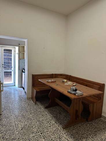 Esta imagen muestra una habitación sencilla, escasamente decorada, con un banco esquinero de madera y una mesa, unos cuantos libros y una cámara sobre la mesa, y suelo de baldosas.