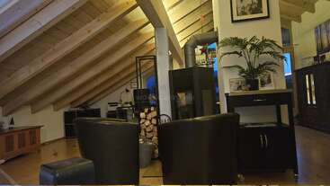 Ce salon mansardé et chaleureux est agrémenté de poutres apparentes, de deux fauteuils, d'un poêle à bois avec une pile de bûches, d'un mobilier moderne, d'une plante en pot, de photos encadrées et d'un éclairage chaleureux.