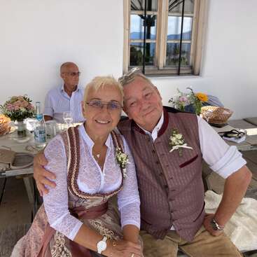 Un couple d'un certain âge, vêtu de costumes traditionnels et arborant des boutonnières fleuries, est assis côte à côte et sourit. Un homme est assis derrière eux à une table décorée.