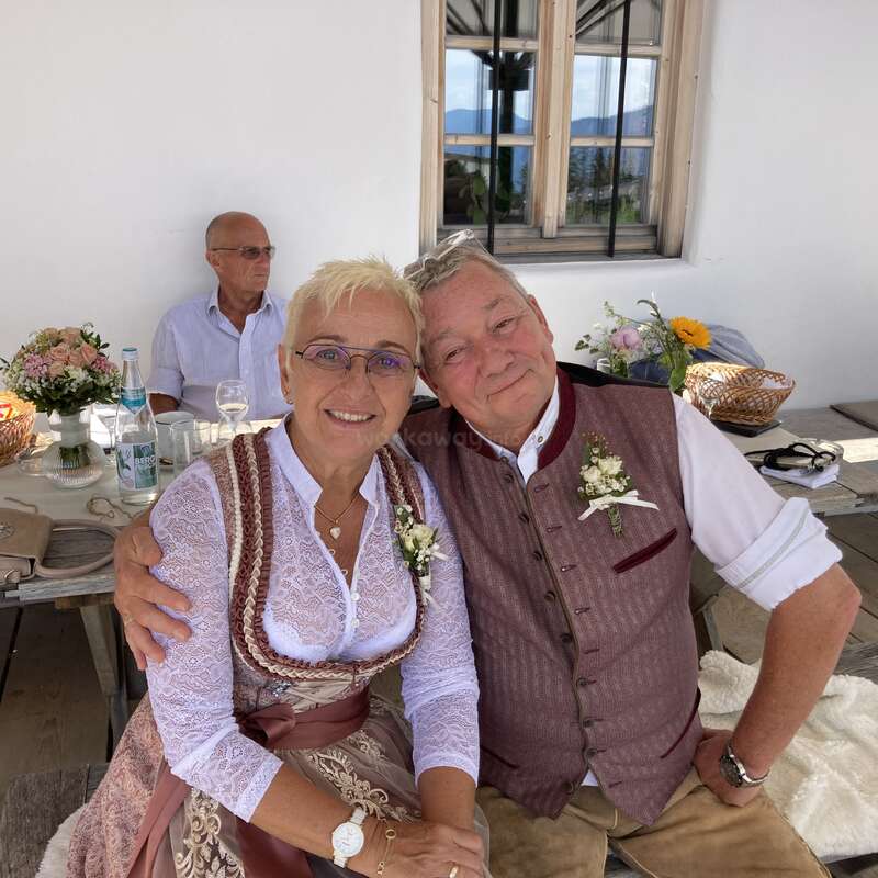 Un couple d'un certain âge, vêtu de costumes traditionnels et arborant des boutonnières fleuries, est assis côte à côte et sourit. Un homme est assis derrière eux à une table décorée.