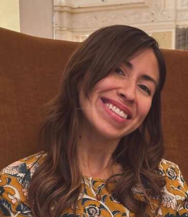 Eine Frau mit langen braunen Haaren sitzt mit einem warmen Lächeln auf einem braunen Stuhl. Sie trägt ein gemustertes gelbes Blumentop. Im Hintergrund sind helle Wände und Dekorationen zu sehen.