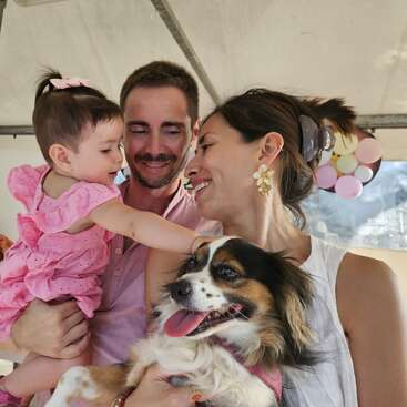 Eine lächelnde dreiköpfige Familie, die einen kleinen Hund hält, genießt einen fröhlichen Moment zusammen. Das Baby greift zärtlich nach dem Hund und strahlt dabei Glück und Liebe aus.