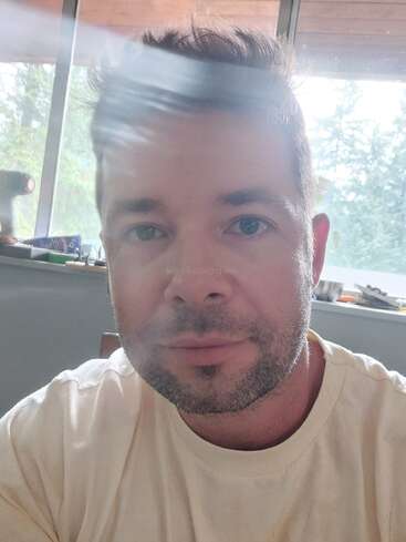 Un homme aux cheveux bruns courts et à la barbe naissante se trouve à l'intérieur, vêtu d'une chemise claire. La lumière du soleil crée une traînée lumineuse sur son visage ; une fenêtre en arrière-plan.