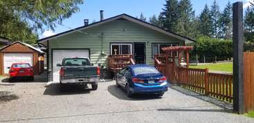 L'image représente une maison à un étage avec un extérieur vert, une porte noire et une voiture bleue garée dans l'allée, entourée d'arbres et d'un ciel clair.
