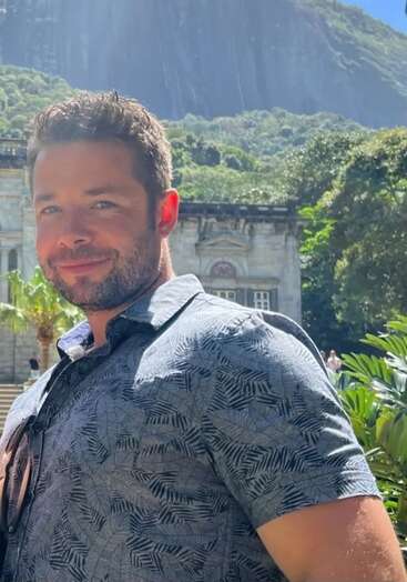 Un homme aux cheveux courts et à la barbe sourit en plein air, à la lumière du soleil. Il porte une chemise à motifs et se tient devant un bâtiment historique et des montagnes.