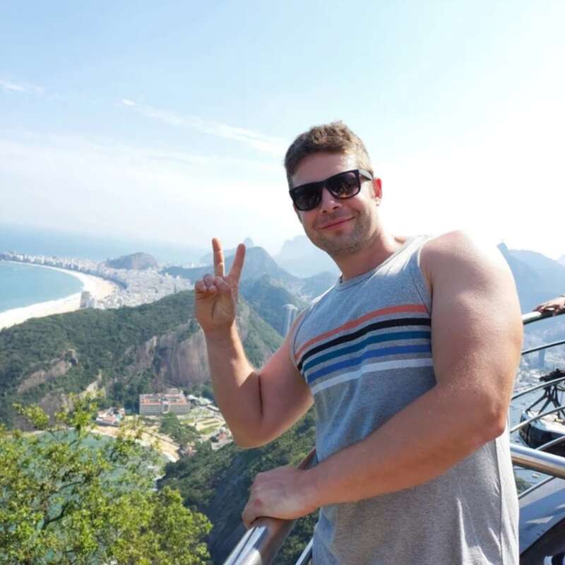 L'image représente un homme debout sur un balcon, surplombant une vue panoramique d'une ville et de montagnes, avec un geste de paix.