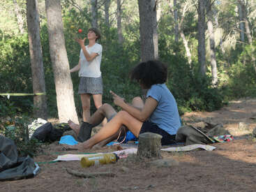 Dos personas se relajan en un bosque. Una está sentada en una manta utilizando un teléfono, mientras que la otra permanece descalza cerca, haciendo malabares con una pelota entre altos árboles.