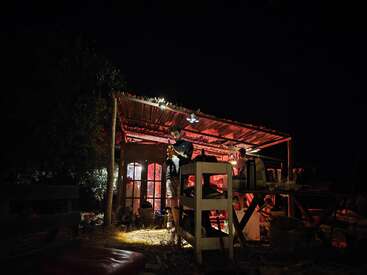Una acogedora escena nocturna al aire libre muestra a un hombre de pie bajo un rústico refugio iluminado en rojo. Varios objetos y personas crean un ambiente cálido, acogedor y animado.