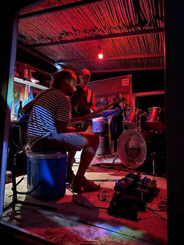 Dos músicos tocan la guitarra y la batería en una pequeña sala con luz roja, rodeados de equipos e instrumentos, creando un ambiente íntimo de jam session.