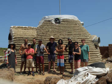 A group of ten people poses in front of a unique, dome-shaped earthbag house under a clear blue sky, suggesting teamwork in sustainable construction.