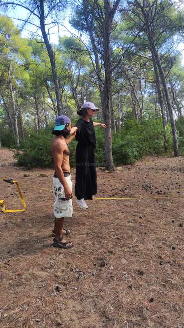 Un hombre sin camiseta ayuda a una mujer con vestido negro y gorra a mantener el equilibrio sobre una cuerda de equilibrio en un bosque con árboles altos y piñas esparcidas.