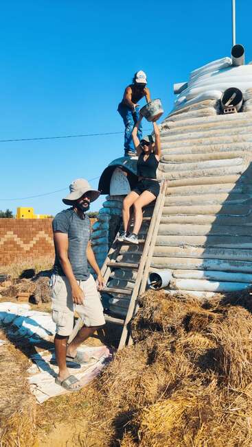 Tres personas trabajan juntas en la construcción de una cúpula de sacos terreros. Uno está de pie en una escalera, otro maneja un cubo y el tercero está de pie debajo, bajo un cielo despejado.