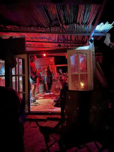 Una banda toca en un pequeño local rústico con techo abierto y una iluminación roja. Los músicos tocan la guitarra y la batería mientras el público observa desde el exterior.