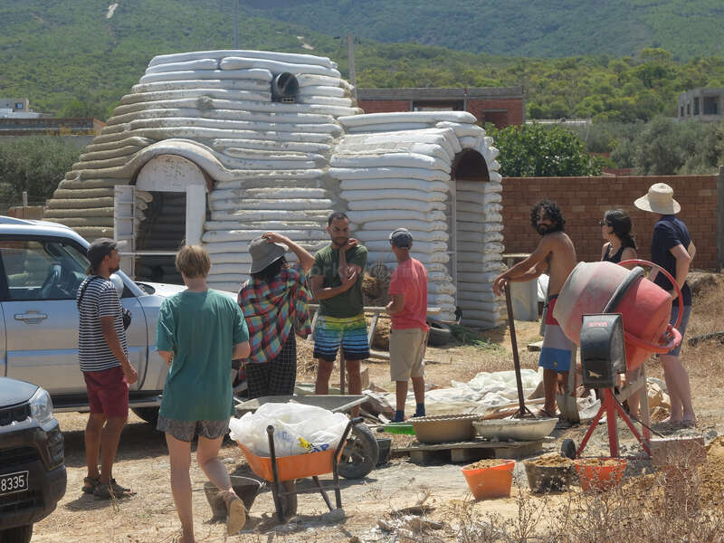 Un grupo de personas está reunido frente a una casa de sacos de tierra en forma de cúpula, aparentemente trabajando en un proyecto de construcción con herramientas, una carretilla y una hormigonera.