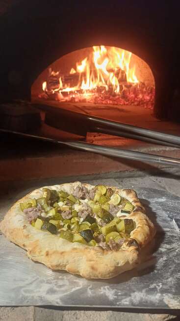 A delicious homemade pizza topped with sausage and zucchini is ready to be baked in a traditional wood-fired oven with flames glowing warmly in the background.