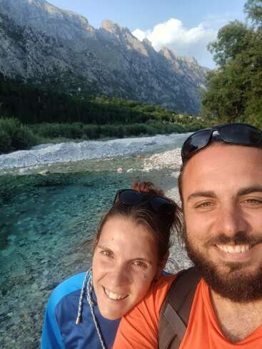 A smiling couple takes a selfie near a clear, shallow river with rocky banks. Majestic mountains and lush greenery form a breathtaking natural backdrop under a sunny sky.