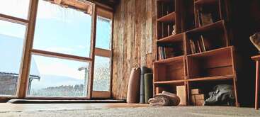 Uma aconchegante sala de ioga ou meditação com janelas amplas, paredes de madeira, estantes de livros, tapetes enrolados, cobertores dobrados e uma luz natural tranquila entrando pela janela, com vista para montanhas nevadas.