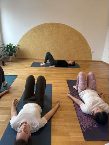 Three people are lying on yoga mats in a serene studio, practicing relaxation or meditation. The room has wooden floors, a yellow mural, and a potted plant.