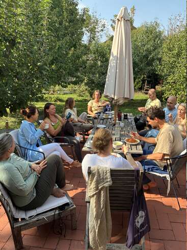 A group of people sit around an outdoor table, enjoying food, drinks, and conversation in a lush garden setting under sunny, clear skies with tall trees.