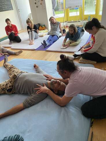 A group sits on mats, attentively taking notes while observing a hands-on demonstration. One person lies on the floor as another gently positions their head and shoulders.
