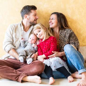 A family of four sits together on a couch, smiling warmly. The father holds a baby, while the mother embraces their young daughter in red. Cozy moment.