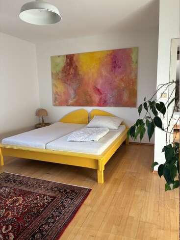 Bright bedroom with a yellow bedframe, abstract colorful painting above bed, white walls, wooden floor, leafy plant, vintage lamp, and a decorative red rug.