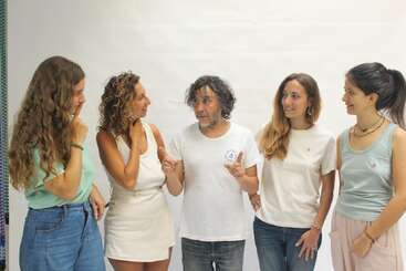 Cinco personas posan juntas ante un fondo blanco, sonriendo e interactuando cordialmente. Cuatro mujeres y un hombre aparecen relajados, amigables y vestidos de manera informal, aparentemente disfrutando de la compañía mutua.
