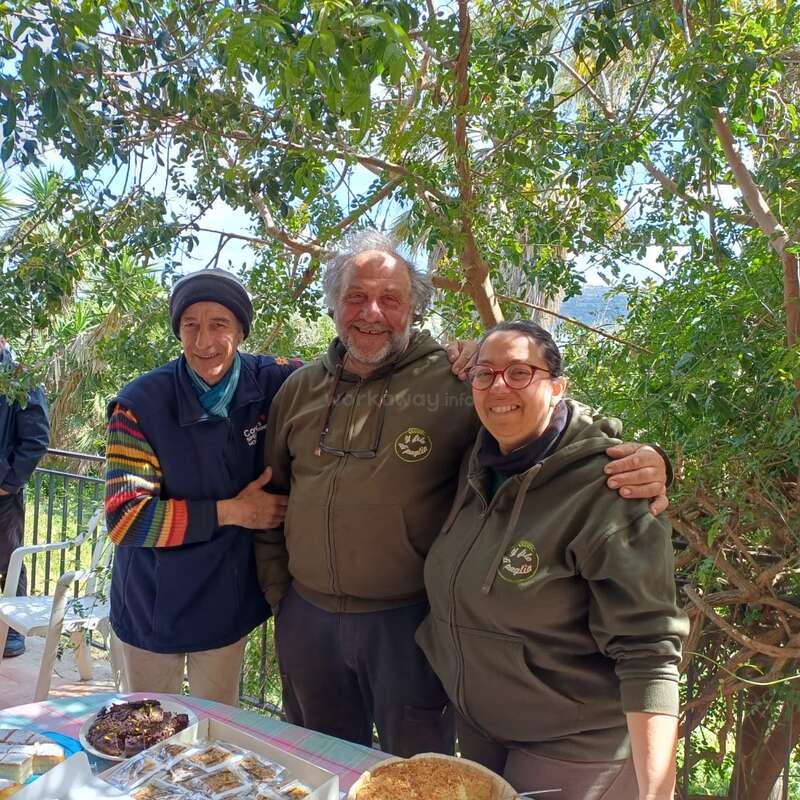Tres personas sonrientes están juntas al aire libre, bajo unos árboles frondosos, disfrutando de la compañía mutua. En la mesa que tienen delante hay una variedad de platos y postres caseros.