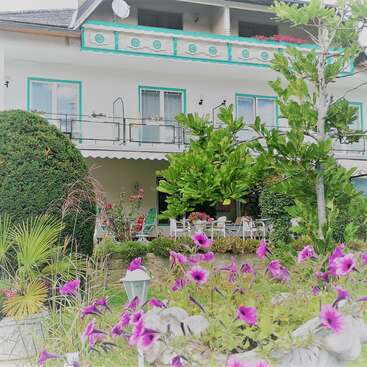 La imagen muestra un sereno edificio blanco con adornos verdes, con balcón y patio, rodeado de frondosos árboles y vibrantes flores moradas en primer plano.