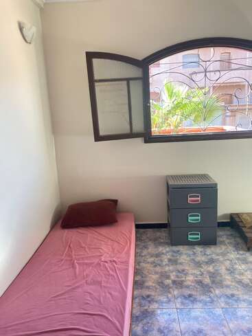 Ein einfaches Zimmer mit einem Einzelbett, das mit einem rosa Laken bedeckt ist, einem roten Kissen, einer Plastikschublade, einem offenen Fenster und einem gefliesten Boden. Pflanzen draußen.