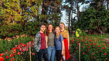 Vier Menschen stehen eng beieinander und lächeln in einem üppigen Garten voller leuchtend roter Blumen. Hohe grüne Bäume bilden den Hintergrund und schaffen eine warme, fröhliche Atmosphäre.