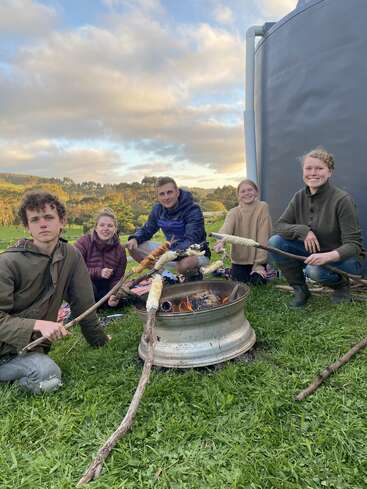 Fünf junge Leute sitzen im Gras um eine behelfsmäßige Feuerstelle und rösten Essen auf Stöcken. Sie sind im Freien, lächeln und genießen den schönen, bewölkten Himmel und die grüne Landschaft.