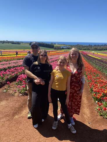 Vier Menschen stehen lächelnd in einem farbenfrohen Tulpenfeld unter einem klaren blauen Himmel, mit Reihen von leuchtenden Blumen und dem Meer im Hintergrund.