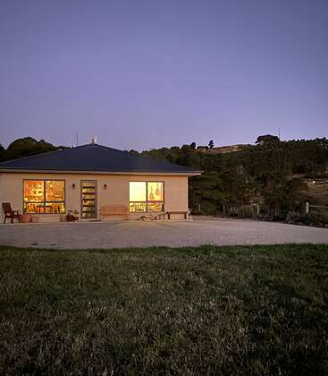 Ein kleines, gemütliches Haus, das von innen warm beleuchtet ist, steht allein in der Abenddämmerung. Umgeben von Gras und Hügeln wirkt es friedlich und einladend gegen den Abendhimmel.