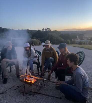 Fünf Menschen sitzen in der Abenddämmerung um ein kleines Lagerfeuer und rösten Essen auf Stöcken. Sie tragen legere, warme Kleidung und Mützen und genießen einen friedlichen Abend im Freien.