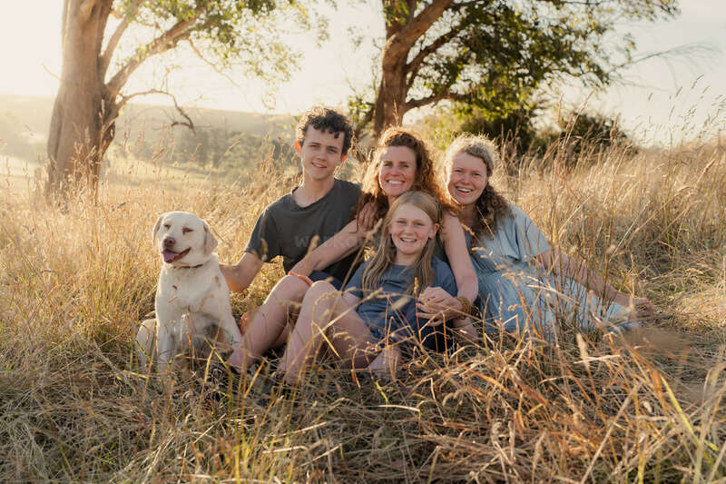 Das Bild zeigt eine Gruppe von Menschen, wahrscheinlich eine Familie, die für ein Foto mit einem Hund vor einer Kulisse aus Bäumen und Büschen posiert, die eine natürliche Umgebung darstellt.