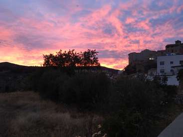 L'image représente une scène sereine de coucher de soleil avec un ciel d'un rose éclatant, des arbres, des bâtiments et des collines en arrière-plan, le tout dans une atmosphère nuageuse.