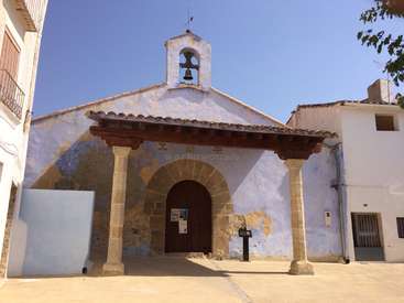 L'image représente une petite église blanche avec un clocher, une porte voûtée et des colonnes de pierre, le tout sur fond de ciel bleu clair.