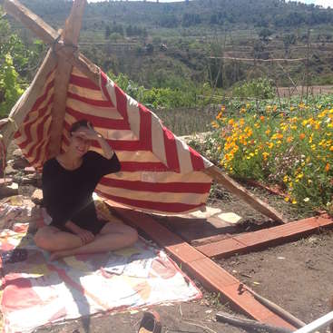 L'image représente une femme assise sur une couverture colorée devant une tente de fortune, entourée d'un jardin aux fleurs éclatantes et d'un paysage pittoresque en arrière-plan.
