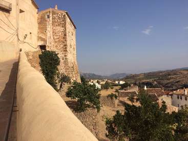 L'image représente une scène de village sereine, avec un bâtiment en pierre à toit plat, entouré d'une végétation luxuriante et de collines ondulantes sous un ciel bleu clair.