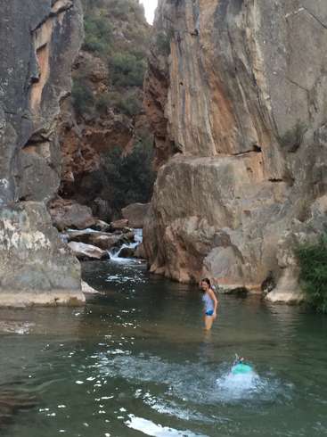 L'image représente un cadre naturel serein, avec une femme debout dans une rivière, entourée de gros rochers et de verdure luxuriante, et une autre personne nageant dans l'eau.