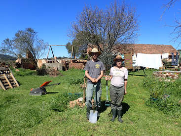 L'image représente un homme et une femme debout dans un espace herbeux, chacun tenant une pelle, avec un bâtiment rustique et des arbres en arrière-plan par une journée ensoleillée.