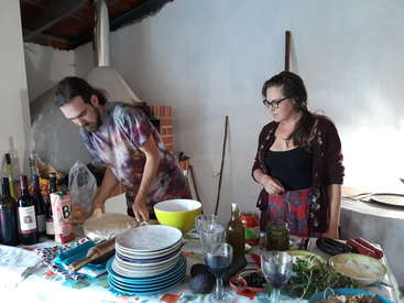 L'image représente un homme et une femme debout derrière une table chargée d'un assortiment d'aliments et de boissons, probablement dans une cuisine ou une salle à manger.
