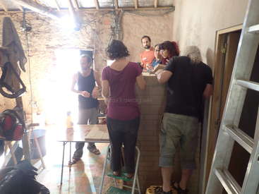 L'image représente un groupe de personnes réunies dans un atelier, avec une échelle, des outils et des équipements visibles, sur fond de mur de briques et de fenêtre lumineuse.