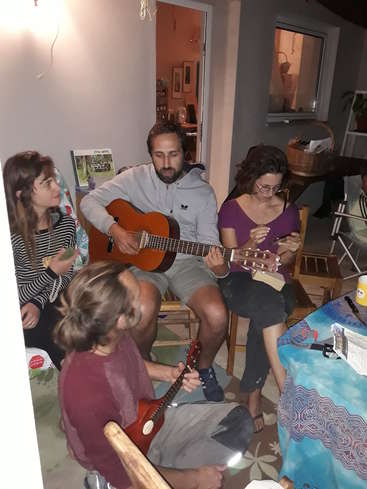 L'image représente un groupe de quatre personnes réunies dans une pièce confortable, deux personnes jouant de la guitare et les autres observant, dans une atmosphère chaleureuse et intime.