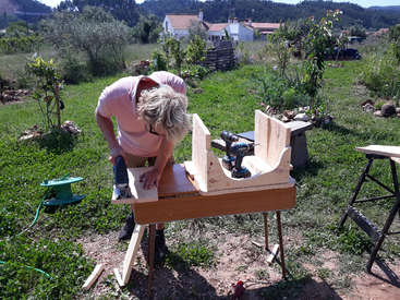 L'image représente un homme âgé travaillant sur un projet en bois dans une arrière-cour, à l'aide d'une perceuse et d'un établi, au milieu d'une végétation luxuriante et d'un cadre rural serein.