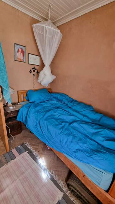 Une chambre confortable avec un lit simple recouvert d'une literie bleue, une moustiquaire suspendue au-dessus, des décorations murales, une petite table de chevet, une lampe, un tapis rayé et des murs couleur pêche.