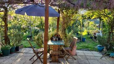 Eine gemütliche Gartenterrasse mit zwei Holzstühlen und einem Tisch unter einem blauen Sonnenschirm. Umgeben von üppigem Grün, Topfpflanzen und durch die Bäume fallendem Sonnenlicht. Friedliche Atmosphäre.