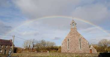 The image depicts a serene scene of a church with a prominent cross, surrounded by a lush green field, with a vibrant rainbow-colored sky and a tranquil body of water in the background.